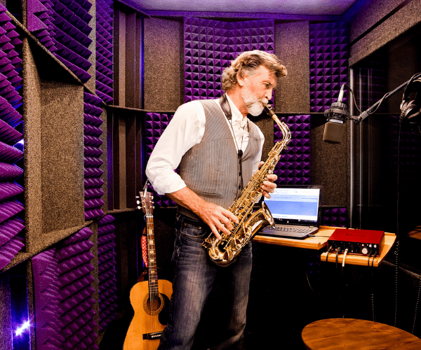 man playing saxophone inside of whisperroom practice booth with a computer and acoustic guitar in the background.