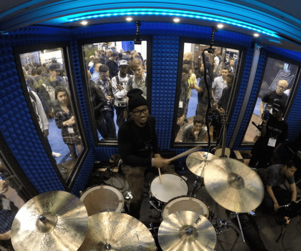 Image of a drummer playing a full kit inside a WhisperRoom drum booth at the NAMM Show. Spectators gather outside, watching the performance through soundproof windows.