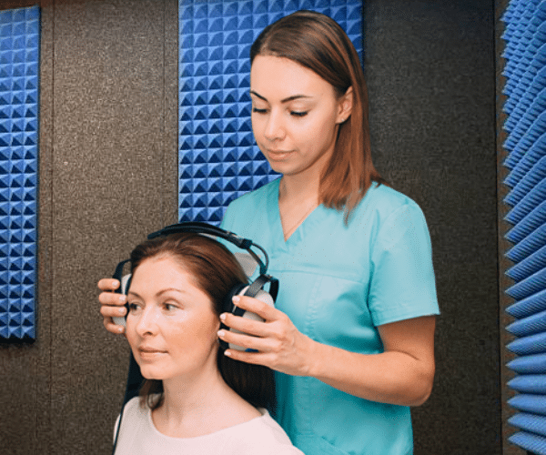 Female audiologist fitting headphones on a woman's head inside a WhisperRoom hearing test booth.