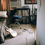 An electric guitar leaning on a bed next to a stack of records