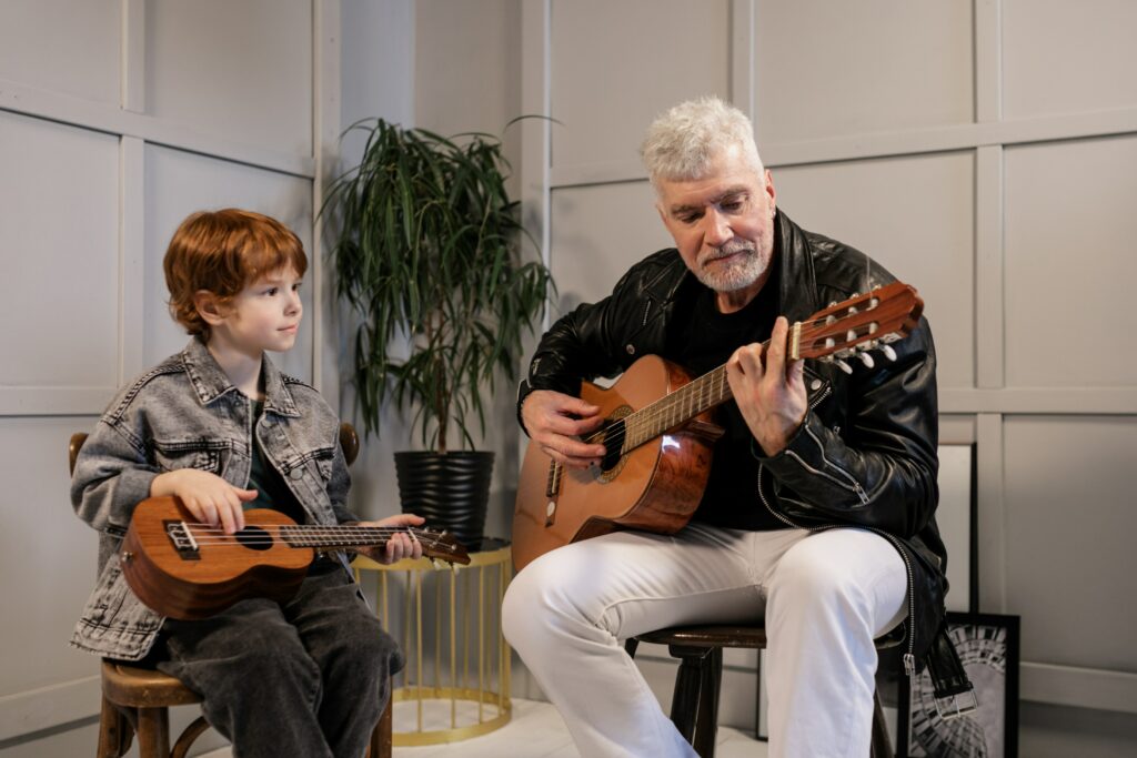 A boy playing ukulele and a man playing a nylon string guitar together in a room