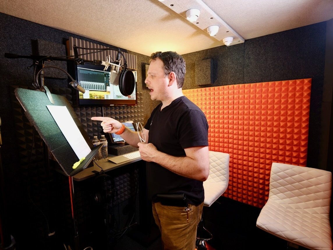 A man recording voice over audio into a microphone while reading a script inside of his WhisperRoom vocal booth.