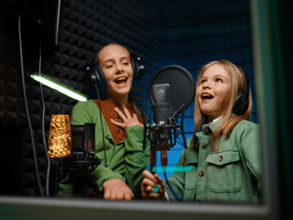 Two girls recording vocals into a condenser mic inside of a soundproof booth for vocals.