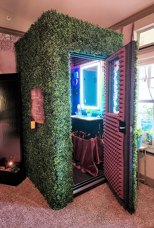 Rachel Fike's WhisperRoom MDL 4848 is shown inside of an apartment with decorative foliage on the outside the sound booth.