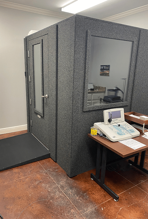 A WhisperRoom Audiology Deluxe Package and hearing test equipment are shown from the side with the door closed inside an audiologist's office.