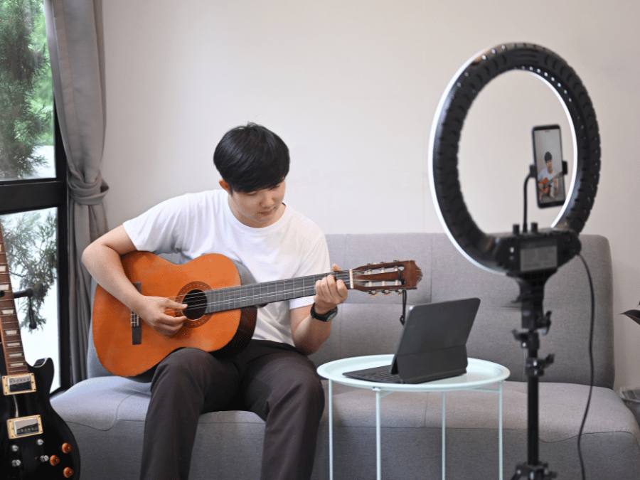 A young man playing guitar on a couch with a live streaming set up for his performance.