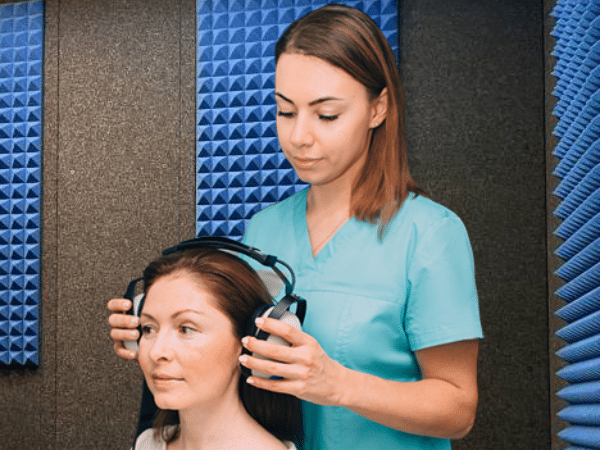 A female audiologist putting headphones on patient to perform a hearing test inside a WhisperRoom audiometric booth.