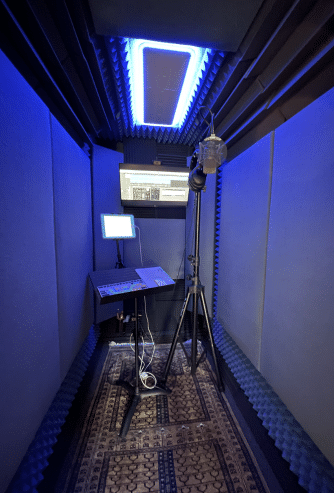 The interior of Todd Stark's WhisperRoom MDL 4872 S shown with acoustic panels on the wall, a computer monitor, and a condenser microphone for recording voice overs.