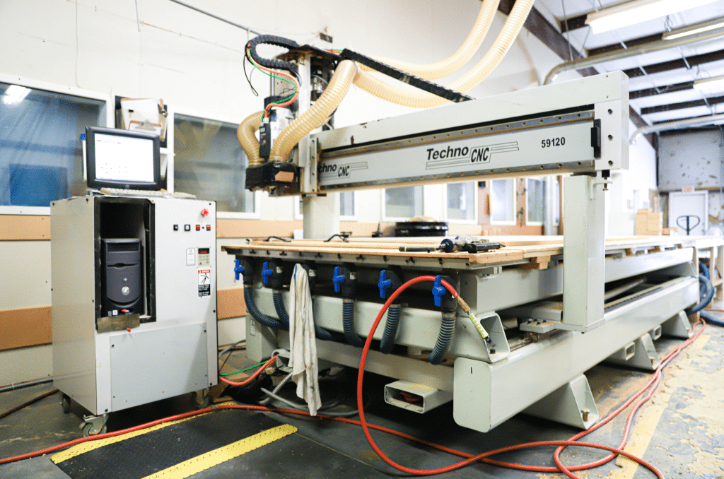 A CNC router in WhisperRoom's woodshop in Morristown, TN, cutting pieces of MDF during the construction process. The router is a large machine with a precision cutting head moving across a large table, carving intricate patterns into the MDF sheets. Sawdust fills the air as the router efficiently transforms the raw material into precise WhisperRoom components.