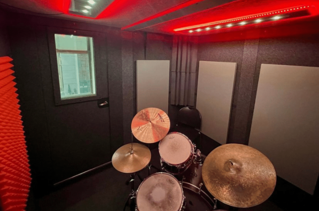 A professionally set up drumkit inside WhisperRoom's Drum Booth Package. The drumkit is positioned in the center of the booth, surrounded by red multi-colored studio lights and acoustic treatment on the walls. This image showcases the ideal drumming environment provided by the Drum Booth Package at Michiko Studios in New York, NY.