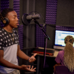 Two college students engaging in a recording session at GCU's Recording Lab. A young man is shown recording vocals into a condenser mic inside a WhisperRoom, while a female student operates the Digital Audio Workstation from a computer outside the booth