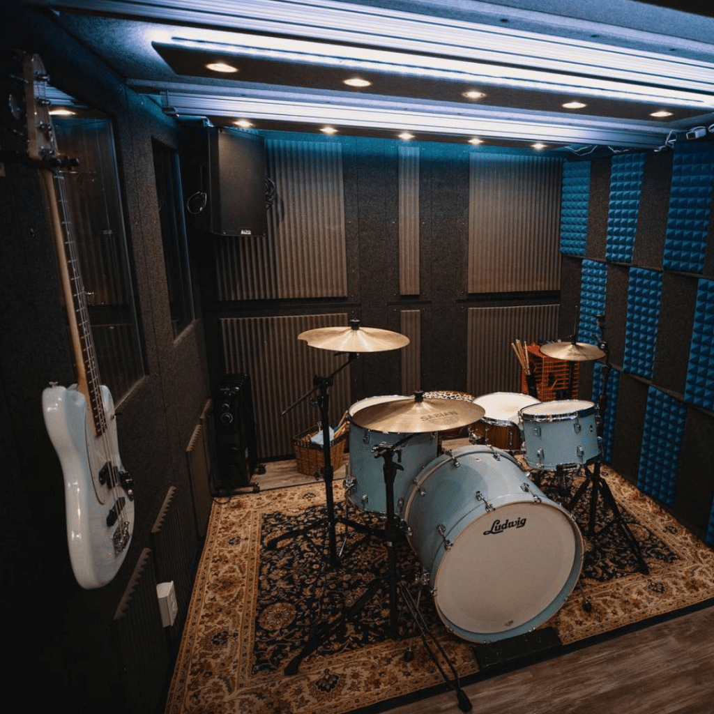 Complete drum kit, bass guitar, and PA speaker set up inside a spacious WhisperRoom practice booth with multiple windows and LED lighting for a premium rehearsal experience.