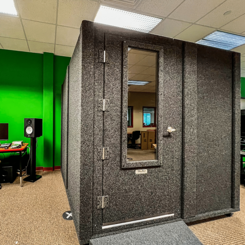 Photograph displaying a 6' x 8' WhisperRoom sound isolation booth placed within a music lab. The booth is adjacent to a recording desk located outside of the booth, illustrating an ideal setup for recording and sound isolation in a music laboratory environment.