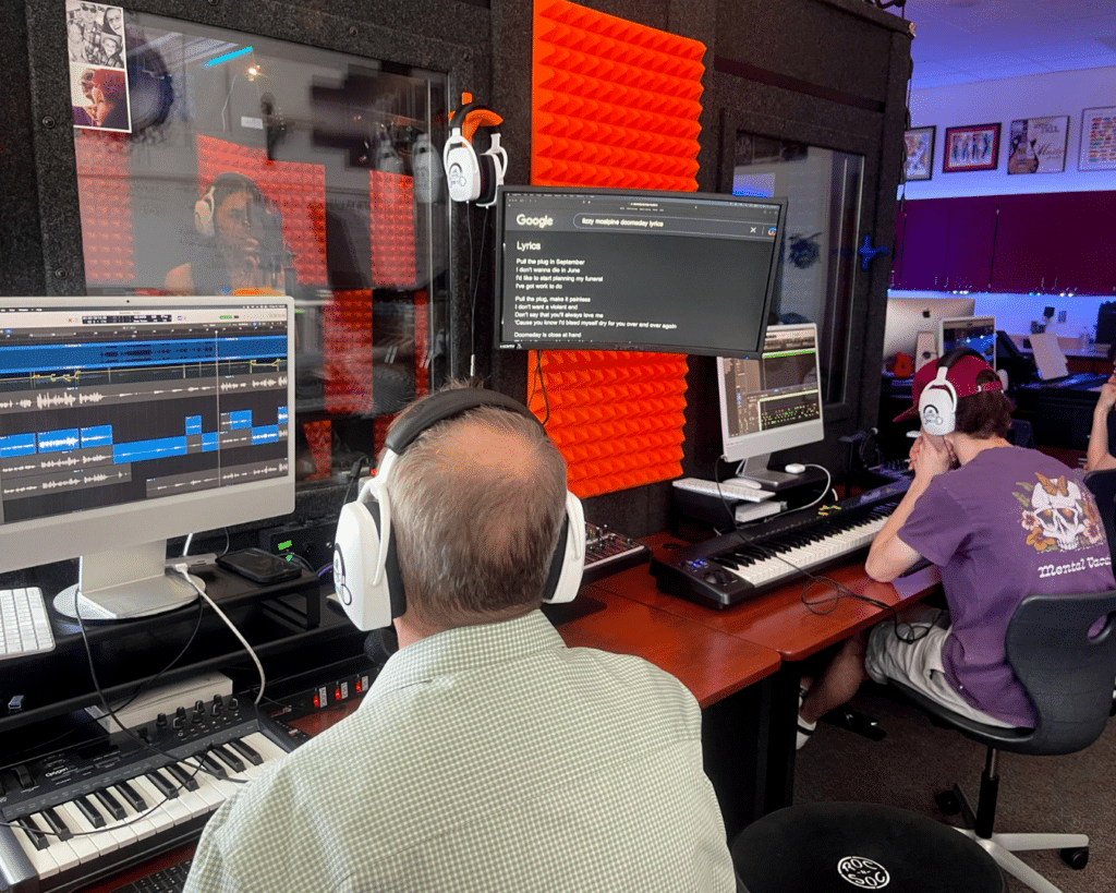 Photograph capturing a music production classroom scene. A teacher and a student are seated at audio editing stations positioned outside of a spacious WhisperRoom sound isolation booth. Inside the booth, a female student is actively recording vocals, while the individuals outside observe the session through a window, creating an interactive and collaborative music learning experience.