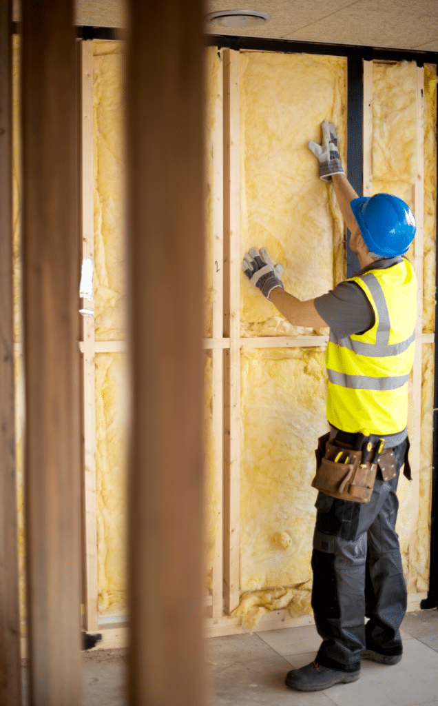 An image of a man wearing a yellow vest and hardhat as he installs insulation between wooden frames. This illustrates the process of soundproofing a home studio, ensuring optimal acoustics and reducing external noise for a professional recording environment.