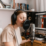 A female musician recording vocals in her home studio, surrounded by professional audio equipment. She wears headphones and sings into a microphone, capturing the essence of a creative workspace. The image highlights the dedication and focus required for success in the music business.